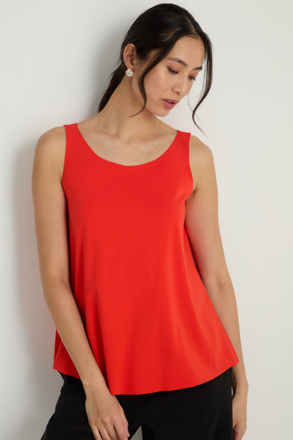 A woman stands against a plain white wall, wearing the Sleeveless Chilli Jersey Top in vibrant red paired with black pants, her head tilted downward to one side.