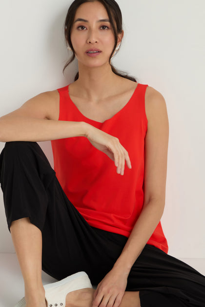 A woman in the Sleeveless Chilli Jersey Top, paired with black pants and white shoes, sits by a white wall with one arm resting on her knee.