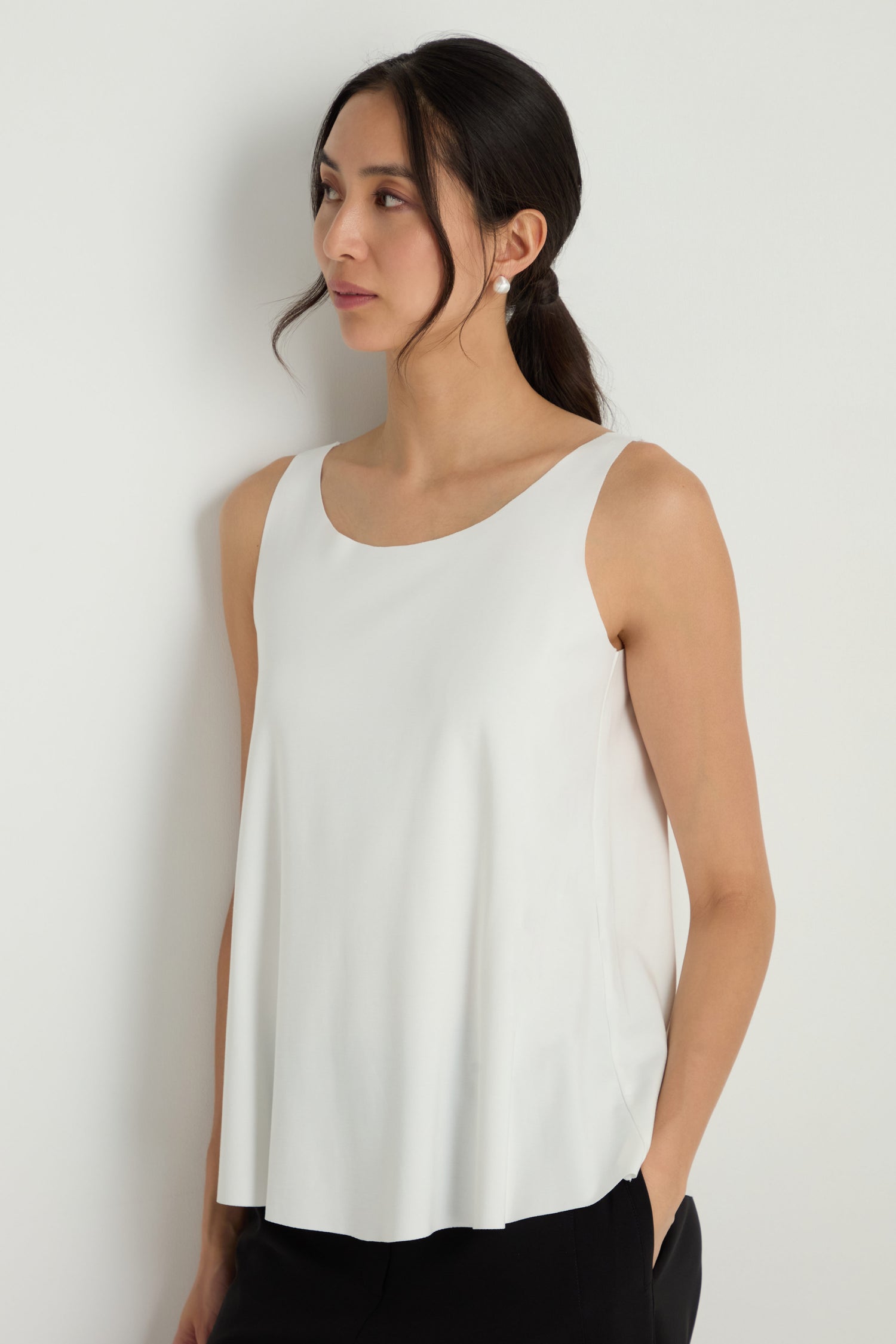 A woman with dark hair in a low ponytail models the Sleeveless White Jersey Top and black pants, standing against a plain white wall with one hand in her pocket.