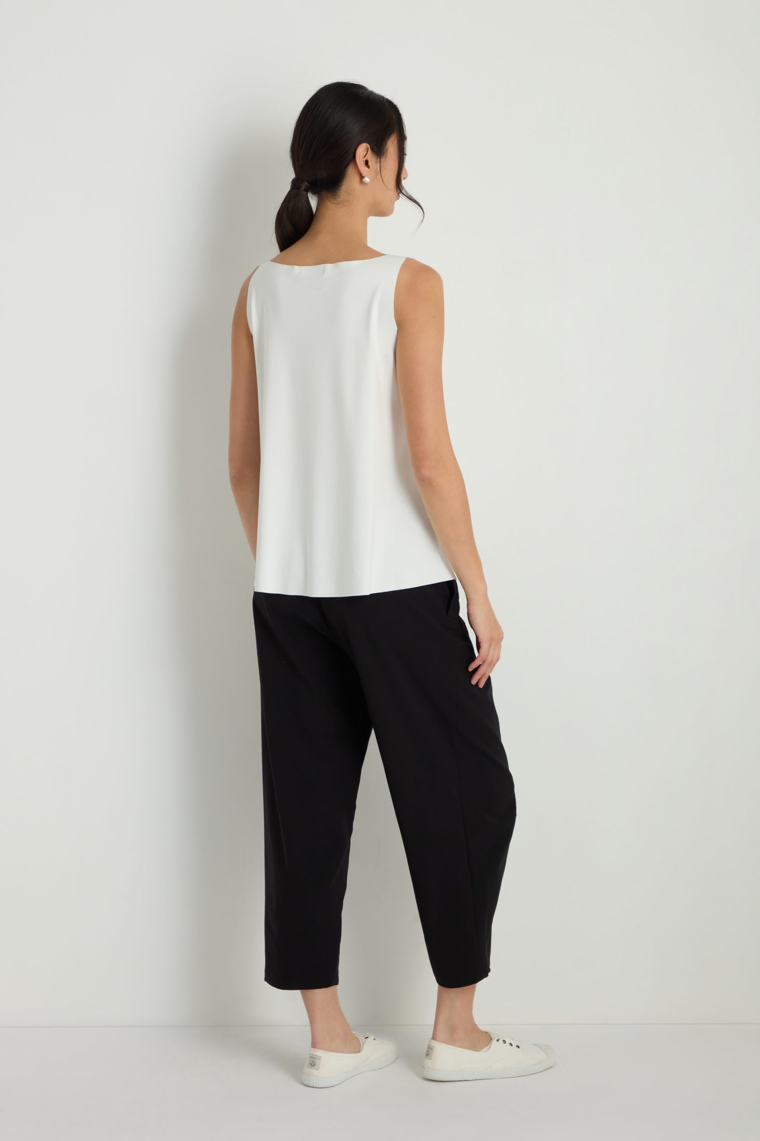 A woman stands against a white wall, facing away, wearing the Sleeveless White Jersey Top, black wide-leg A-line pants, and white sneakers.