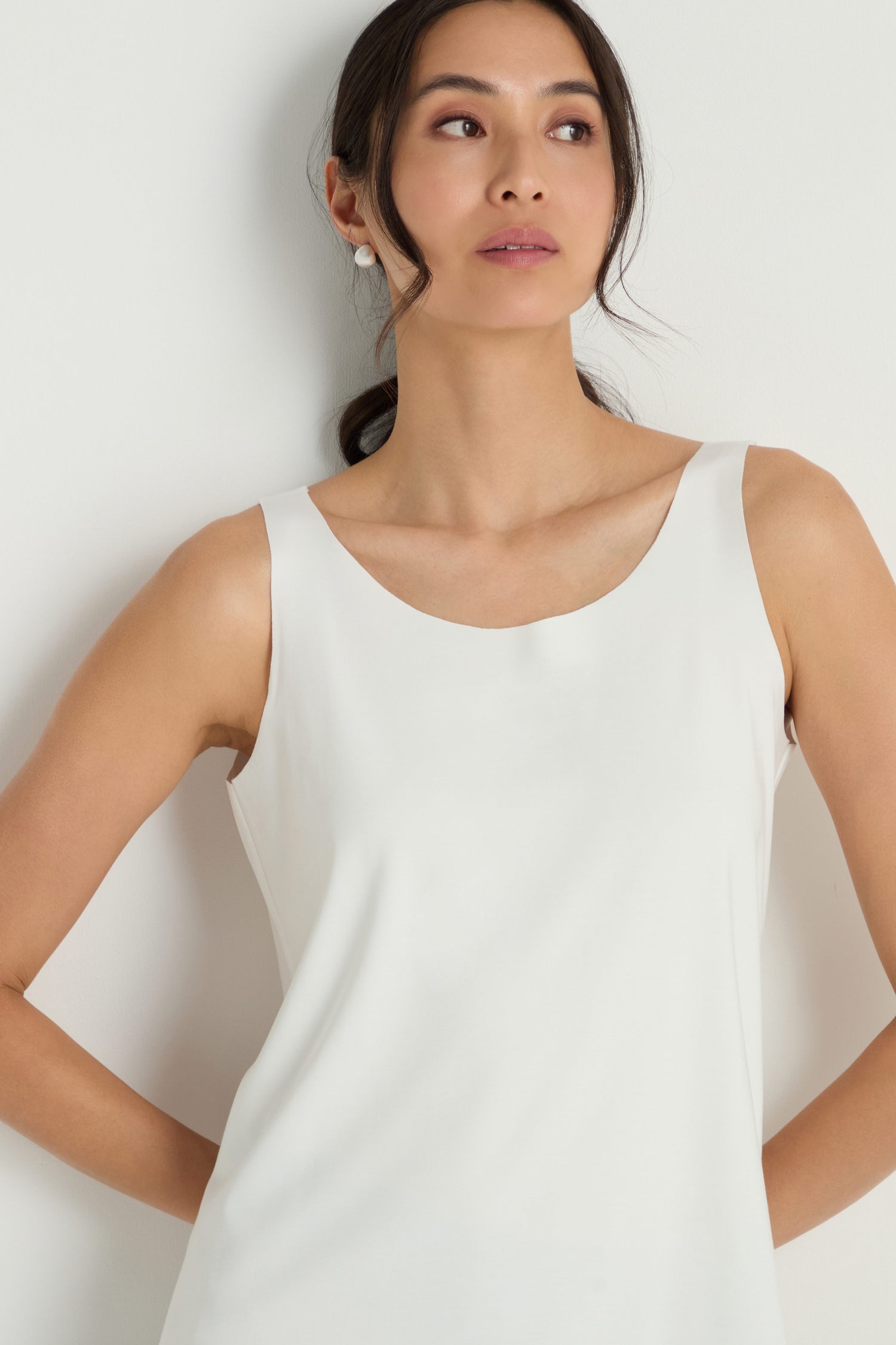 A woman with long dark hair pulled back is wearing the Sleeveless White Jersey Top, standing against a plain white background and looking slightly to the side.