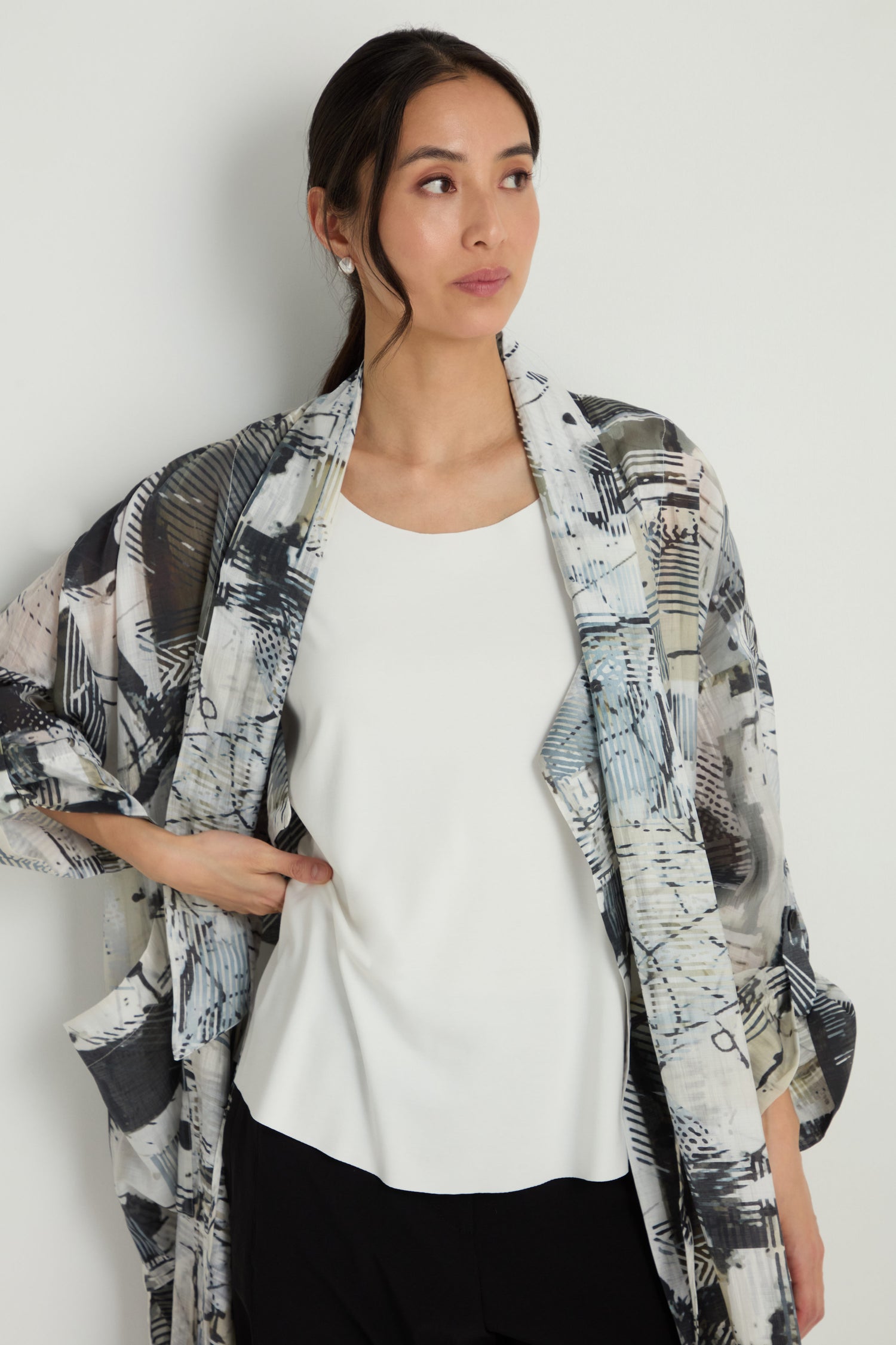 A woman stands against a white wall, wearing the Sleeveless White Jersey Top under a patterned kimono-style jacket and black A-line pants, looking to the side with one hand on her hip.