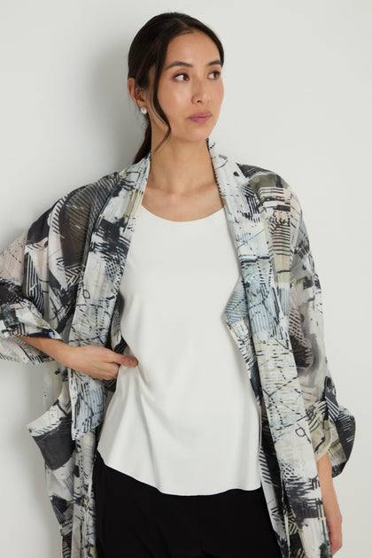 A woman stands against a white wall, wearing the Sleeveless White Jersey Top under a patterned kimono-style jacket and black A-line pants, looking to the side with one hand on her hip.