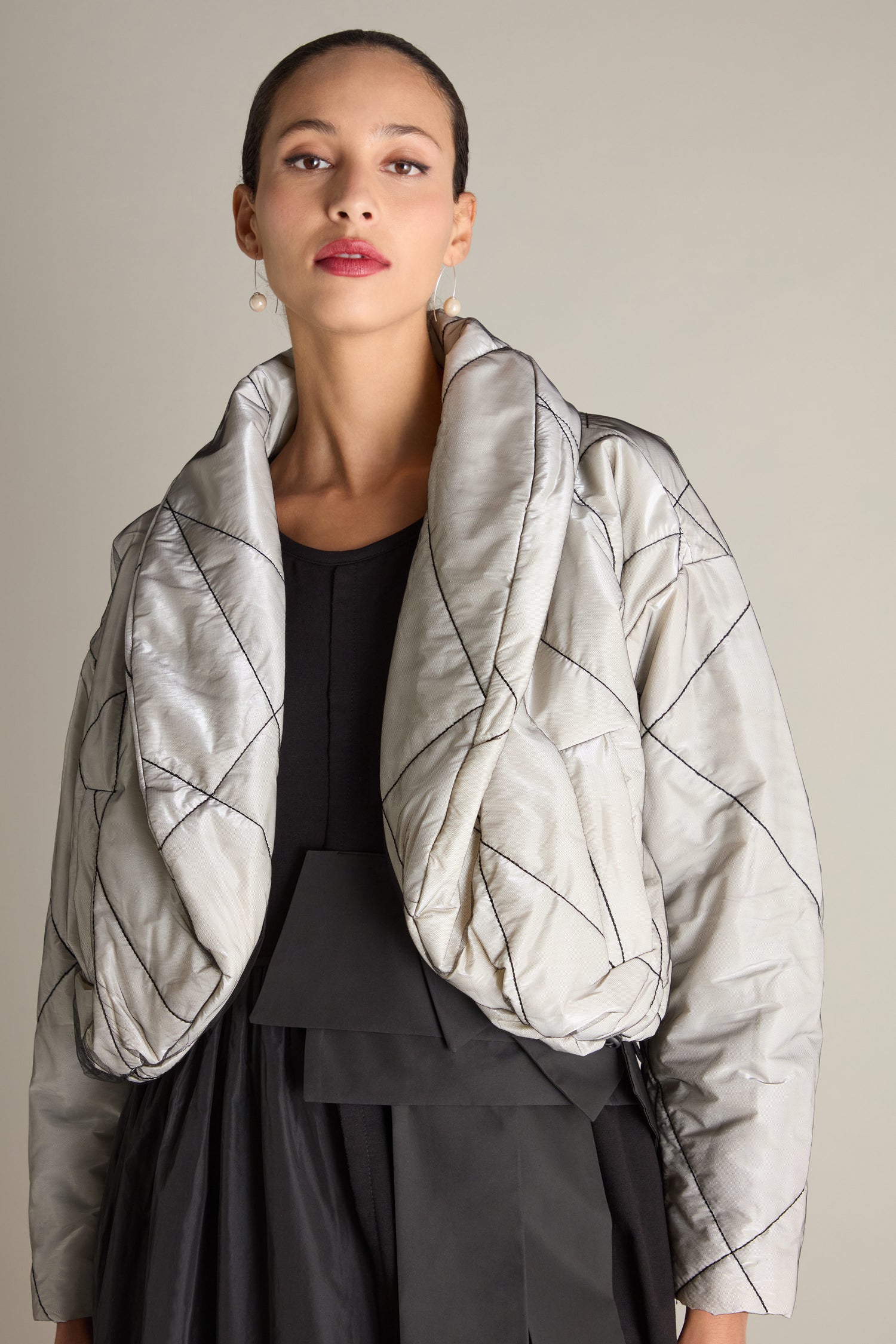 A woman stands against a plain background wearing a black outfit layered with the Short Puffa Evening Jacket, which features a textured, light-colored design with embroidered detailing. She has a neutral expression and wears subtle earrings.