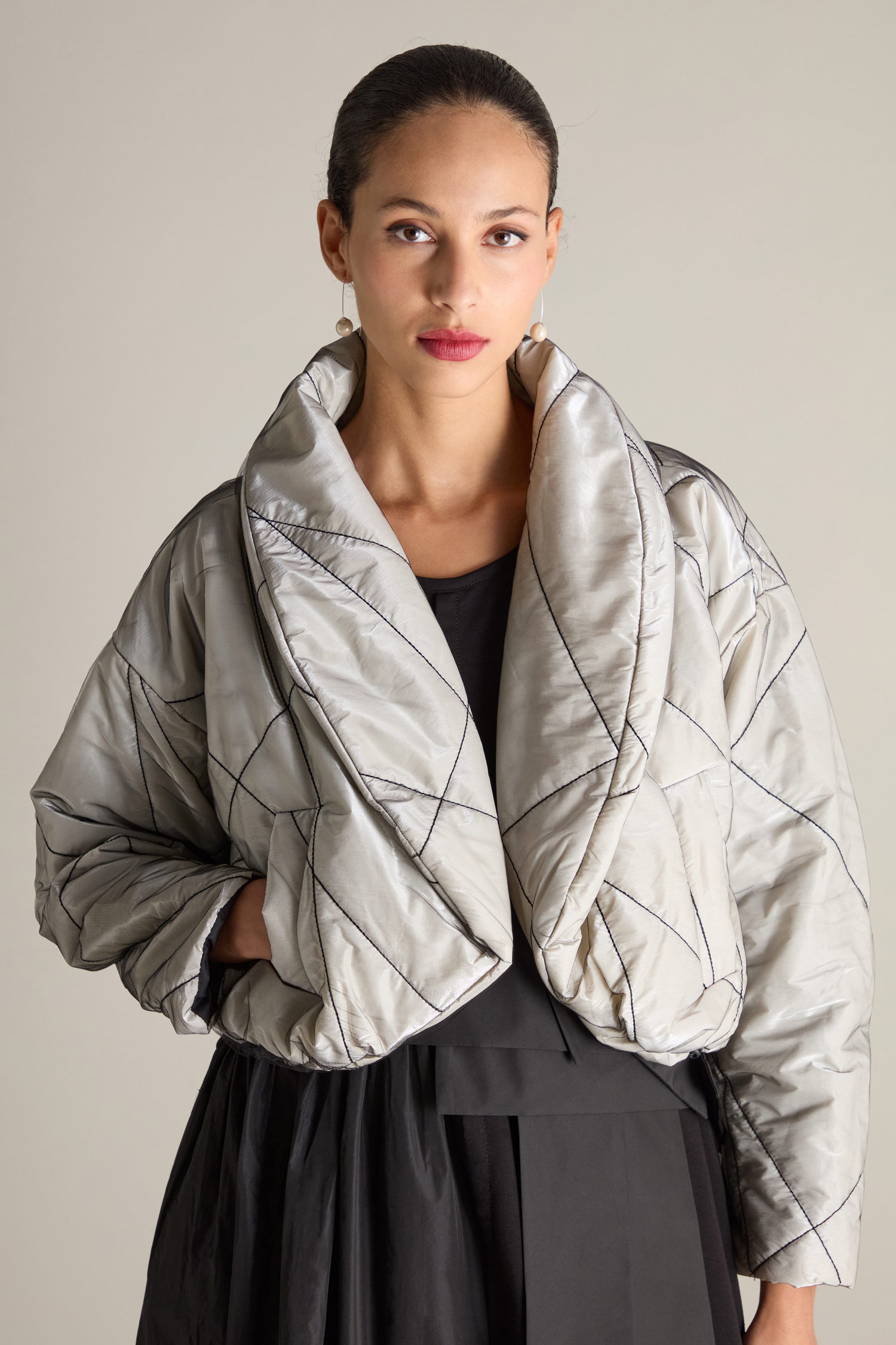 A woman with pulled-back hair is wearing the Short Puffa Evening Jacket, a quilted, metallic silver jacket featuring a large collar and black stitching, over a black outfit. She stands against a plain backdrop, gazing forward.