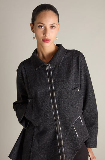 A person with a neutral expression is wearing the Zip Detail Jacket, characterized by its dark color, white stitching, and oversized front pockets, posing against a plain background.