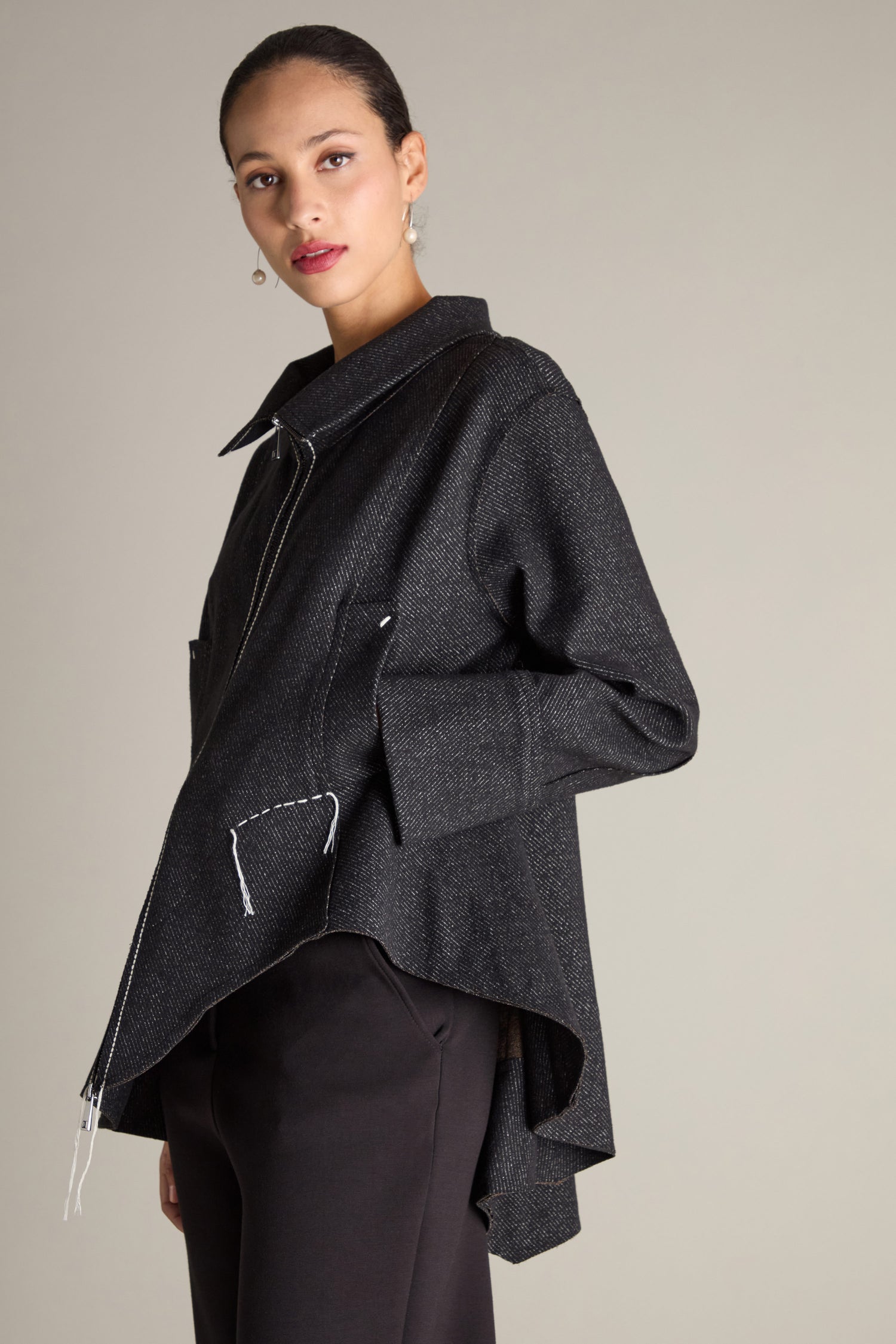 A person wearing the Zip Detail Jacket, which features a dark color, asymmetrical design, oversized front pockets, and visible stitching, stands against a plain background.