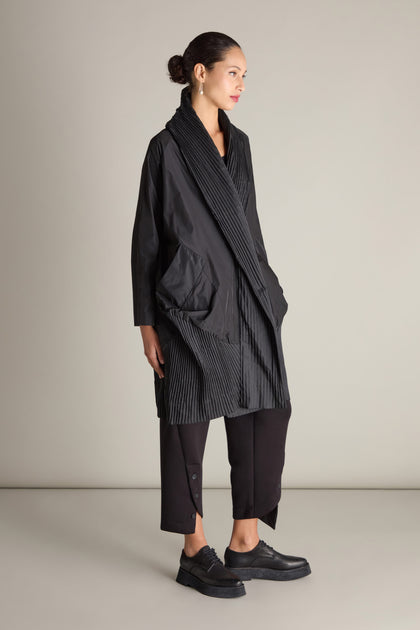 A person stands sideways wearing a black, oversized Rib Detail Evening Coat featuring a textured design, paired with black pants and black shoes. Their hair is styled in a bun, and they are posed against a simple, light background.