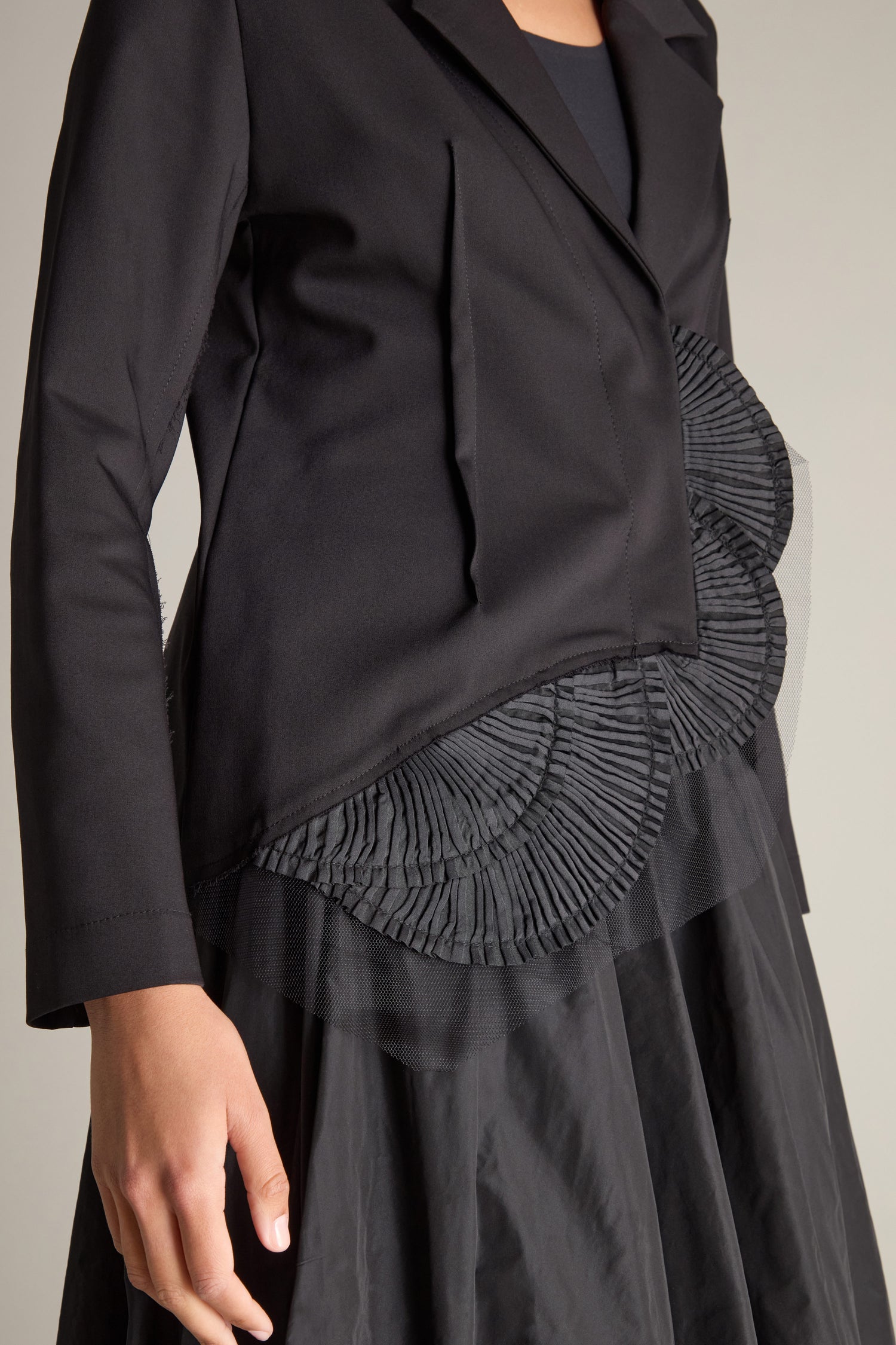 A close-up of a person wearing the Fitted Fan Detail Jacket, showcasing its pleated waist details over a black skirt. The jacket is elegantly designed with long sleeves, a classic lapel collar, and adorned with 3D fan frills applique.