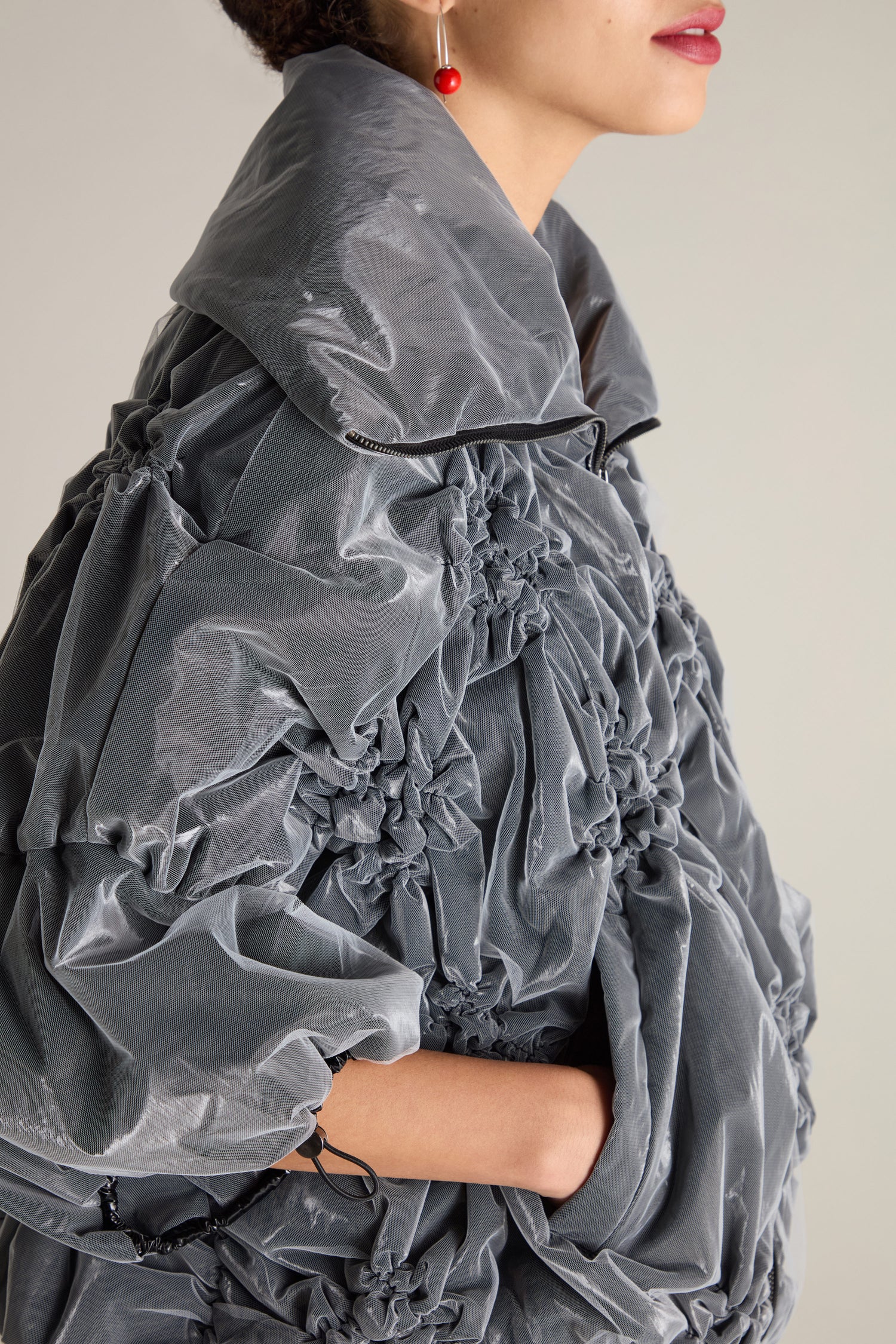 A person wearing a gray Ruched Collar Zip Jacket, showcasing delicate ruched fabric details and drawstring cuffs, paired with a red dangling earring. This modern statement piece stands out as the individual gazes to the right.