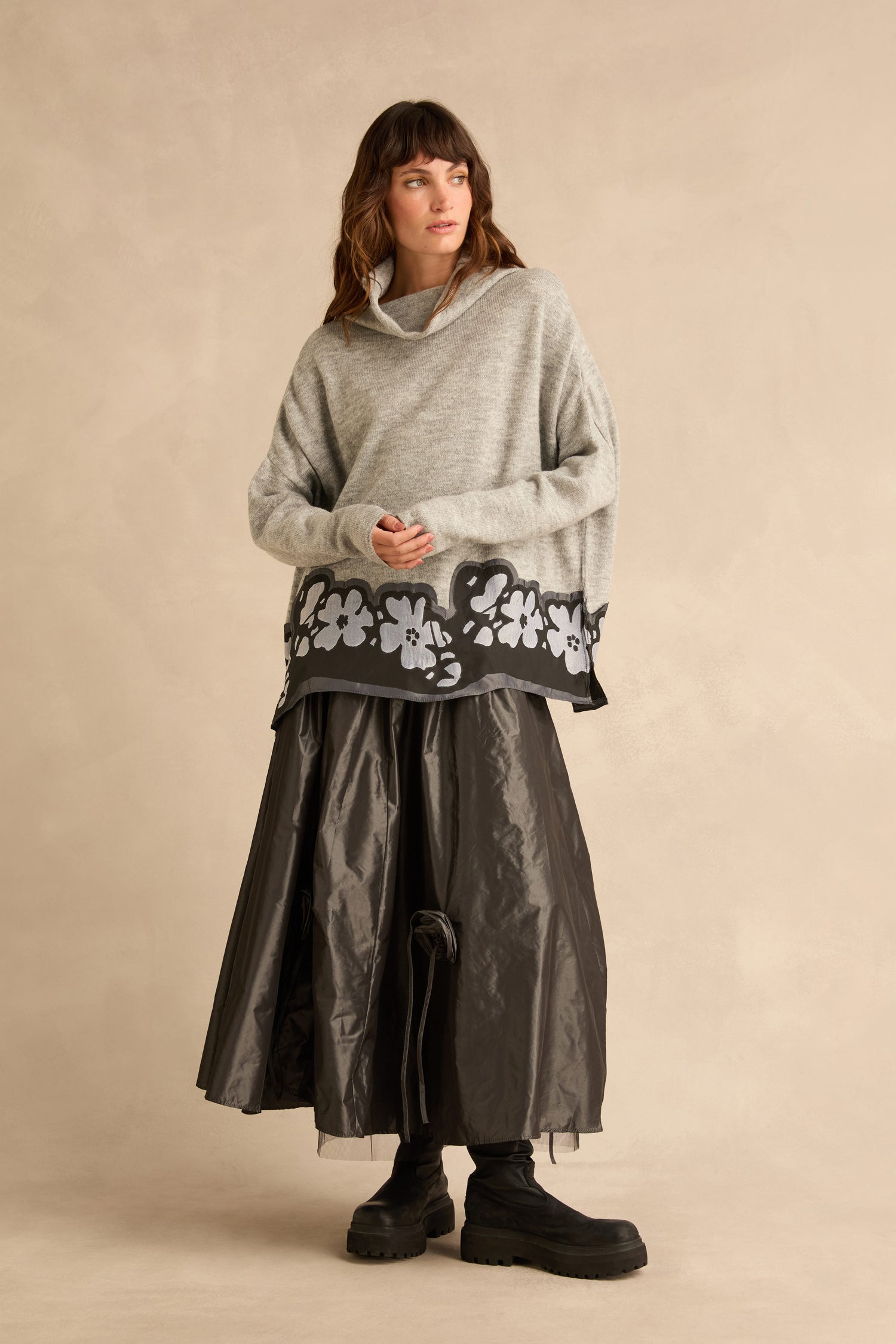 A person wearing a grey Flower Hem Knit sweater with a floral appliqué at the hem, a long dark skirt, and black boots.