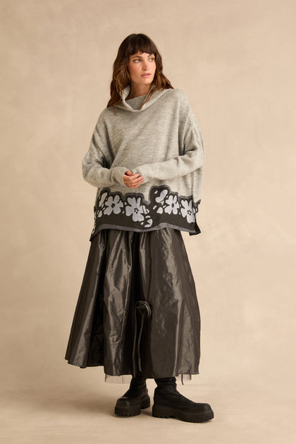 A person wearing a grey Flower Hem Knit sweater with a floral appliqué at the hem, a long dark skirt, and black boots.