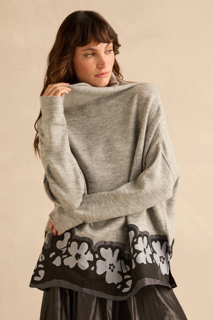 A woman wearing an oversized grey Flower Hem Knit sweater with a black floral appliqué hem.