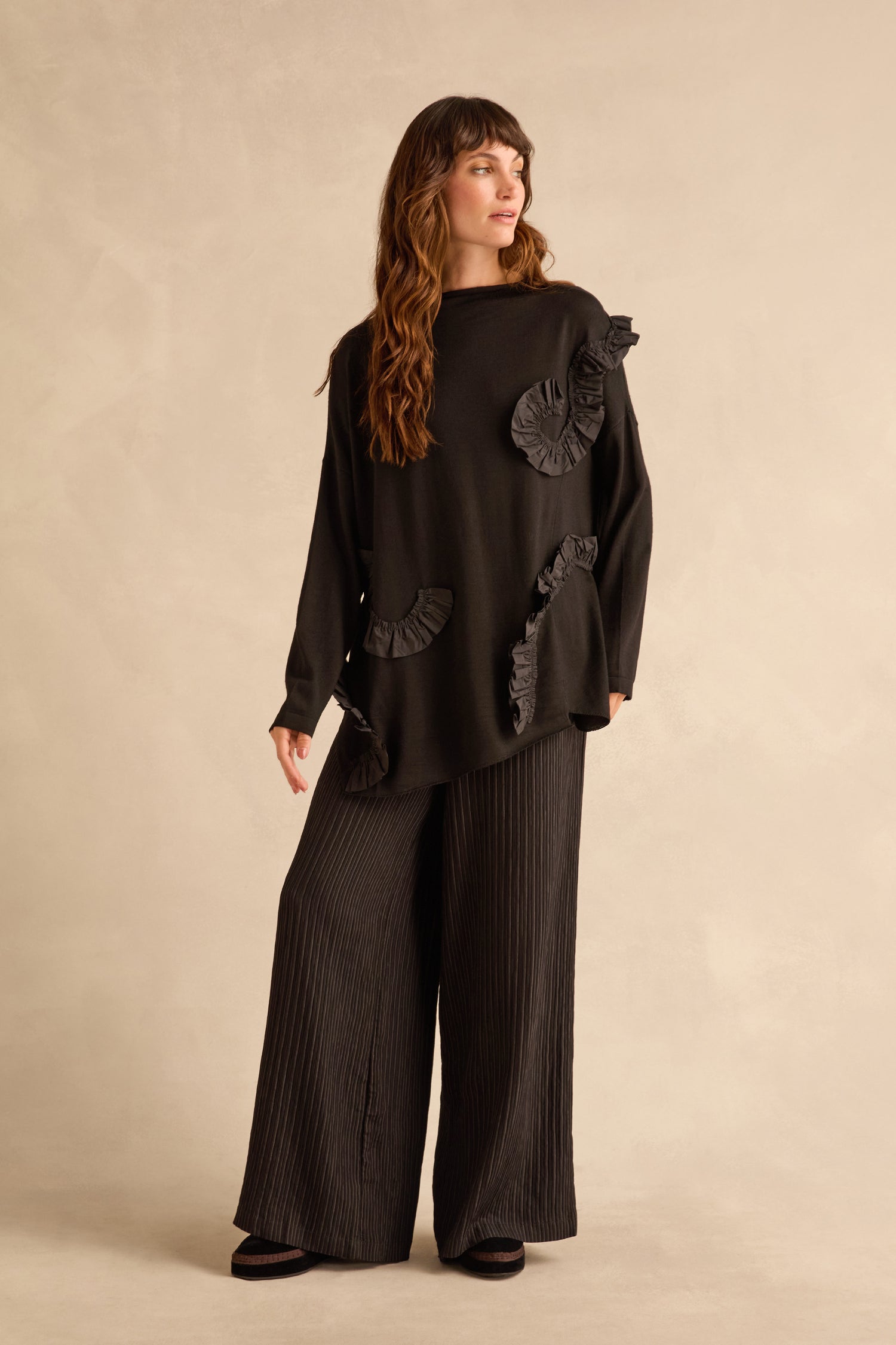 A woman wears a black Taffeta Detail Knit Top with asymmetrical taffeta ruffle appliqué and wide-leg pleated trousers.