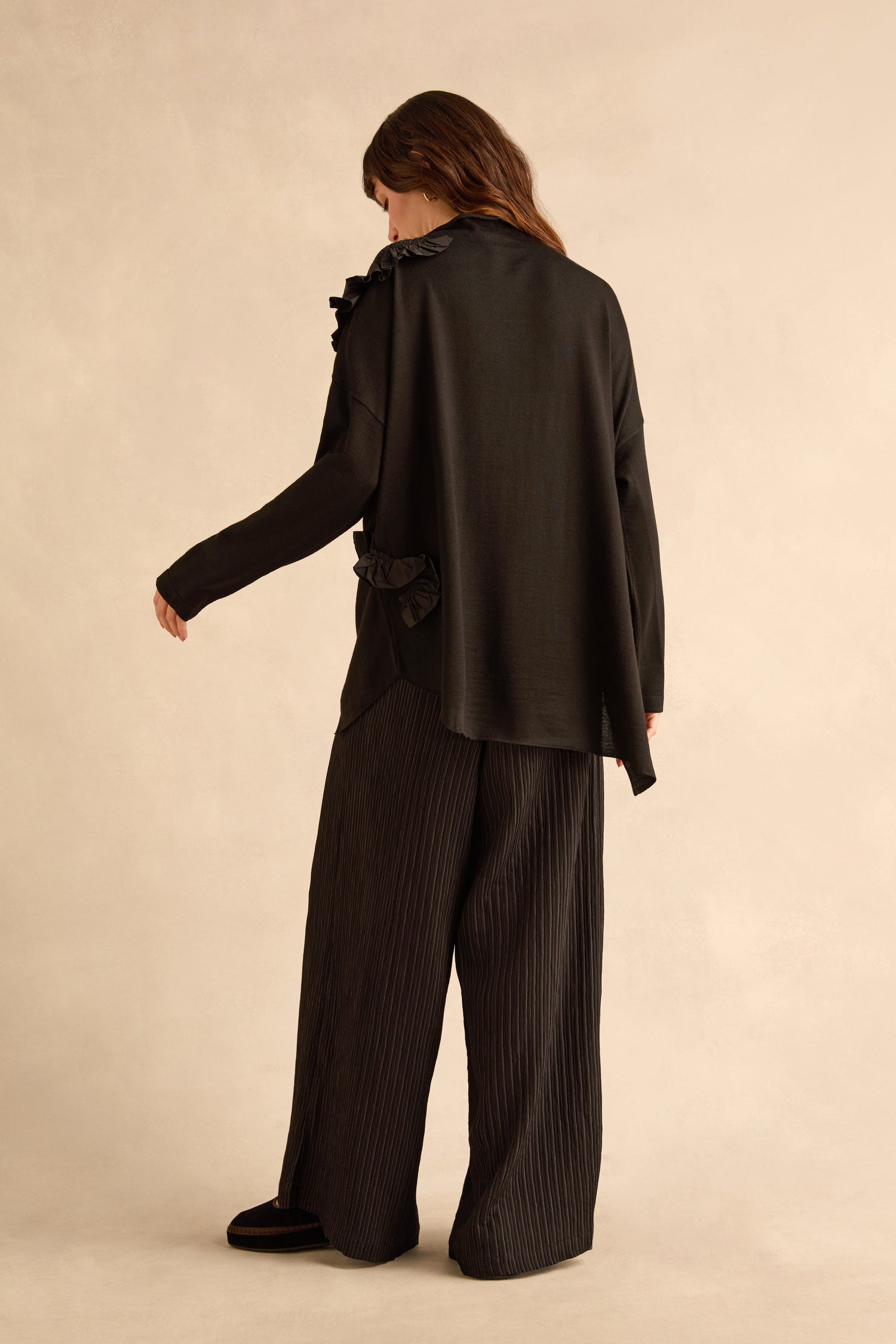 Back view of a person wearing a black Taffeta Detail Knit Top with taffeta ruffle appliqué at the shoulder and hip, paired with wide-leg pleated trousers.