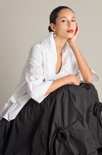 A woman in a white Frill Neck Easy Top featuring 3/4 sleeves and cuffs, paired with a black skirt, poses with her hand resting on her cheek.