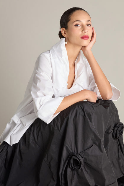 A woman in a white Frill Neck Easy Top featuring 3/4 sleeves and cuffs, paired with a black skirt, poses with her hand resting on her cheek.