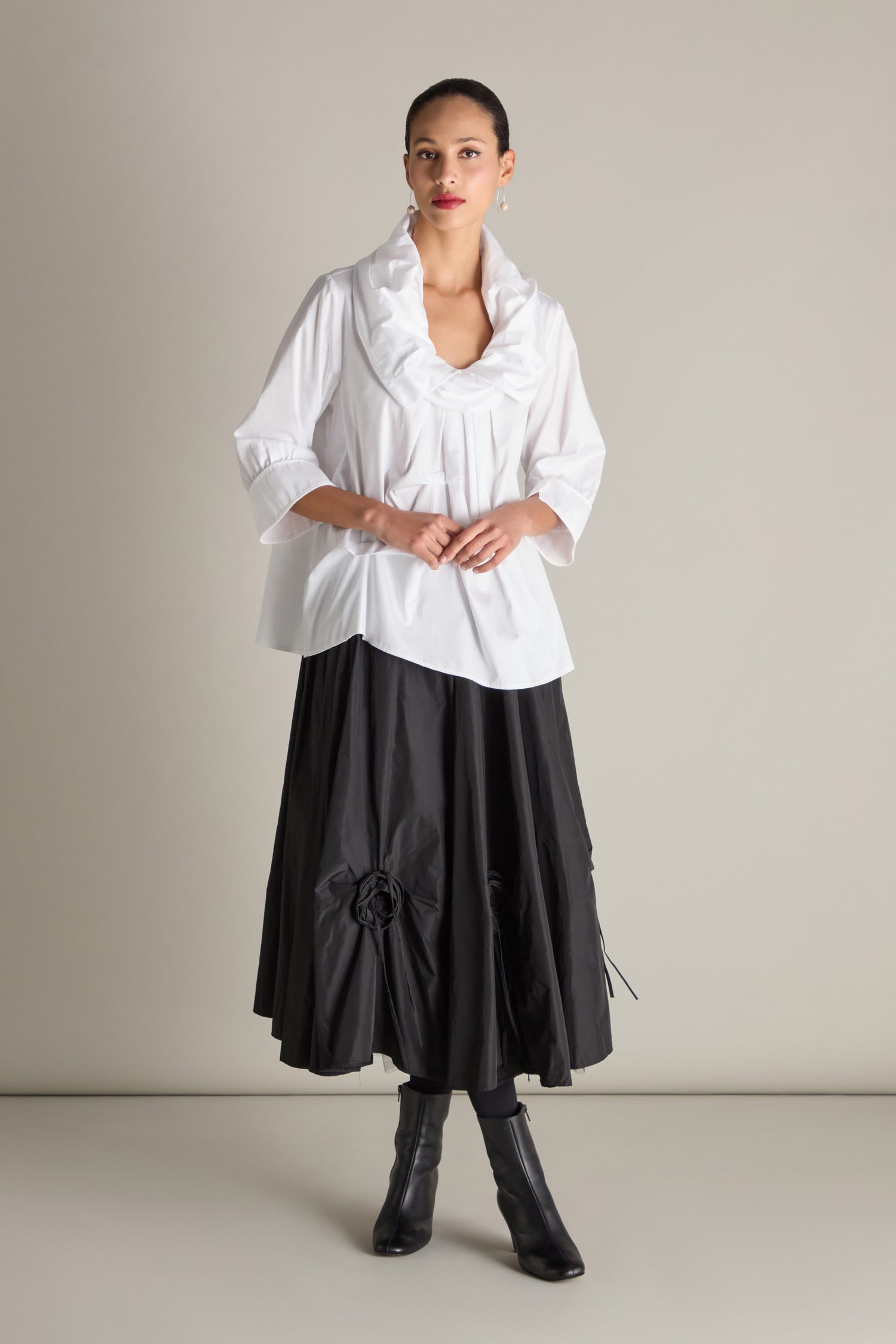 A person stands against a plain background, wearing a Frill Neck Easy Top with 3/4 sleeves and cuffs, paired with a black skirt adorned with floral details, and complemented by black boots.