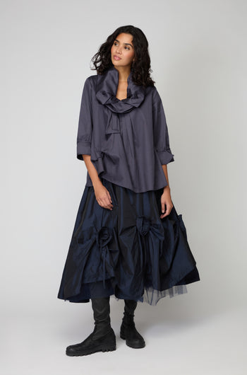 A woman stands against a plain background wearing the Frill Neck Easy Top in dark blue, featuring a ruffled collar and three-quarter sleeves, paired with a navy voluminous skirt with large bows and black boots.
