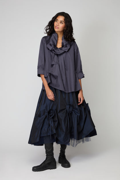 A woman stands against a plain background wearing the Frill Neck Easy Top in dark blue, featuring a ruffled collar and three-quarter sleeves, paired with a navy voluminous skirt with large bows and black boots.