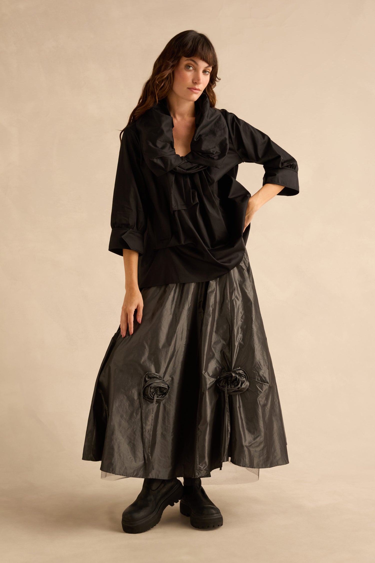 A person wears a Black Frill Neck Easy Top with a relaxed silhouette and large ruffled collar, paired with a dark, full-length skirt with rosette accents and chunky black boots.