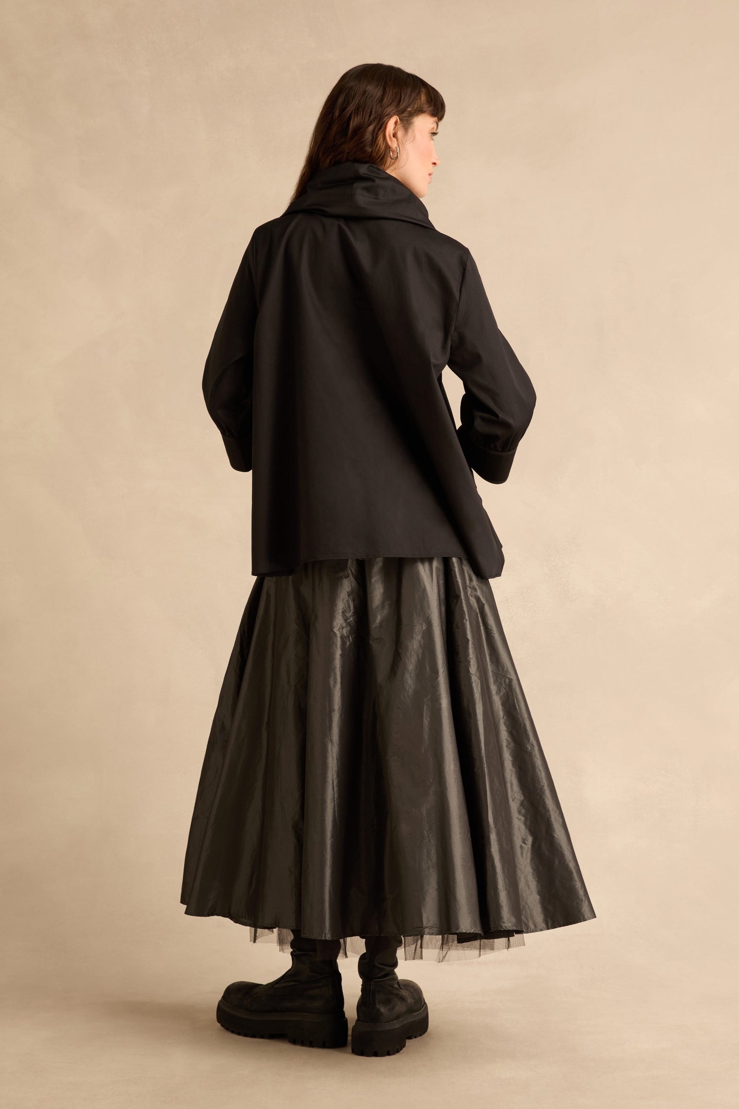 Seen from behind, a person wears a Black Frill Neck Easy Top, a long, full black skirt, and chunky black boots.
