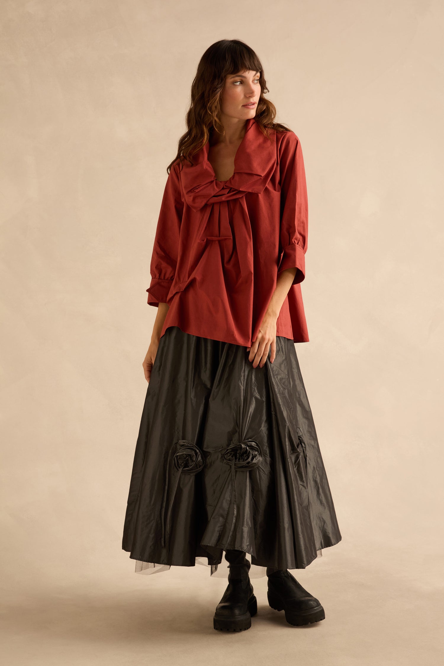 A woman wearing a Red Frill Neck Easy Top with three-quarter sleeves, paired with a long, pleated black skirt featuring circular fabric embellishments and black platform boots.