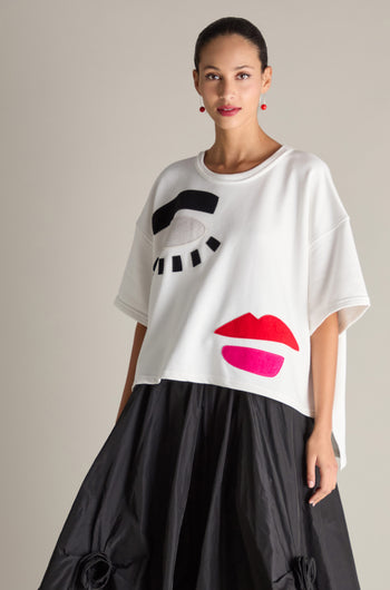 A person is wearing the Eyes And Lips Jersey Top, a white oversized T-shirt embellished with abstract black and red eye and lip designs, paired with a black skirt.