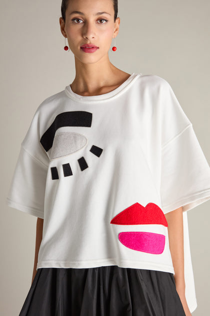 A person wearing the Eyes And Lips Jersey Top, featuring stylized eye and lips graphics, stands against a plain background. Adorned with red earrings, they gaze into the camera, beautifully highlighting the abstract design of the top.