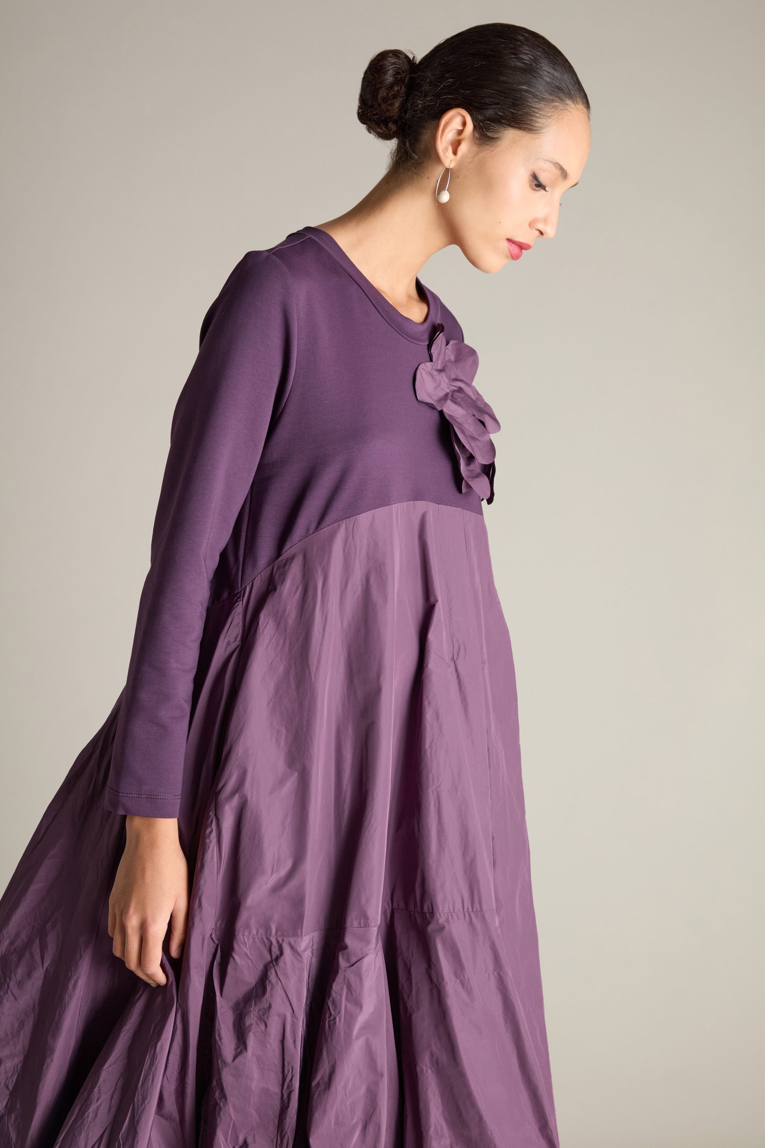 A person elegantly poses against a neutral background, wearing the Jersey Taffeta Flower Dress, a stylish long purple garment with an empire waistline and adorned with a floral accessory on the shoulder.