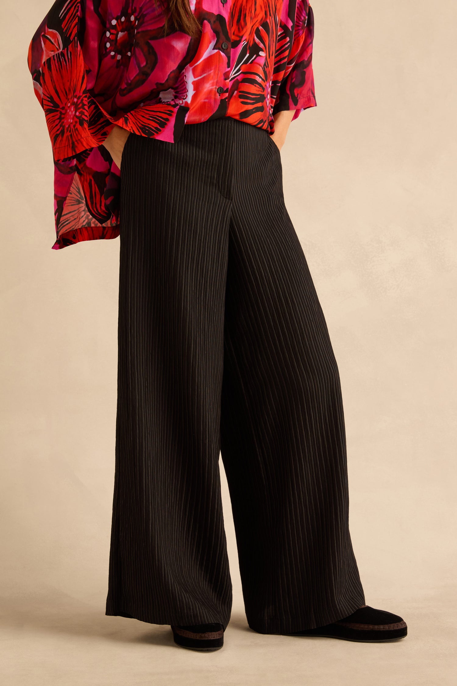 A person wears black Pin Stripe Wide Leg Trousers with a vibrant red and pink floral print button-down shirt.