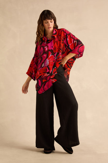 A woman wears a long-sleeved shirt with a bright red and pink floral print tucked into black Pin Stripe Wide Leg Trousers.