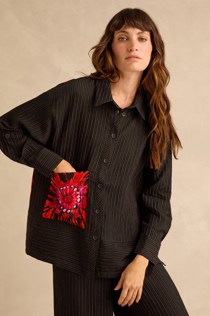 A person wearing a Flora Pin Stripe Shirt, which is a relaxed-fit, black, thin-striped, button-down shirt featuring a bright red and black floral-patterned chest pocket.