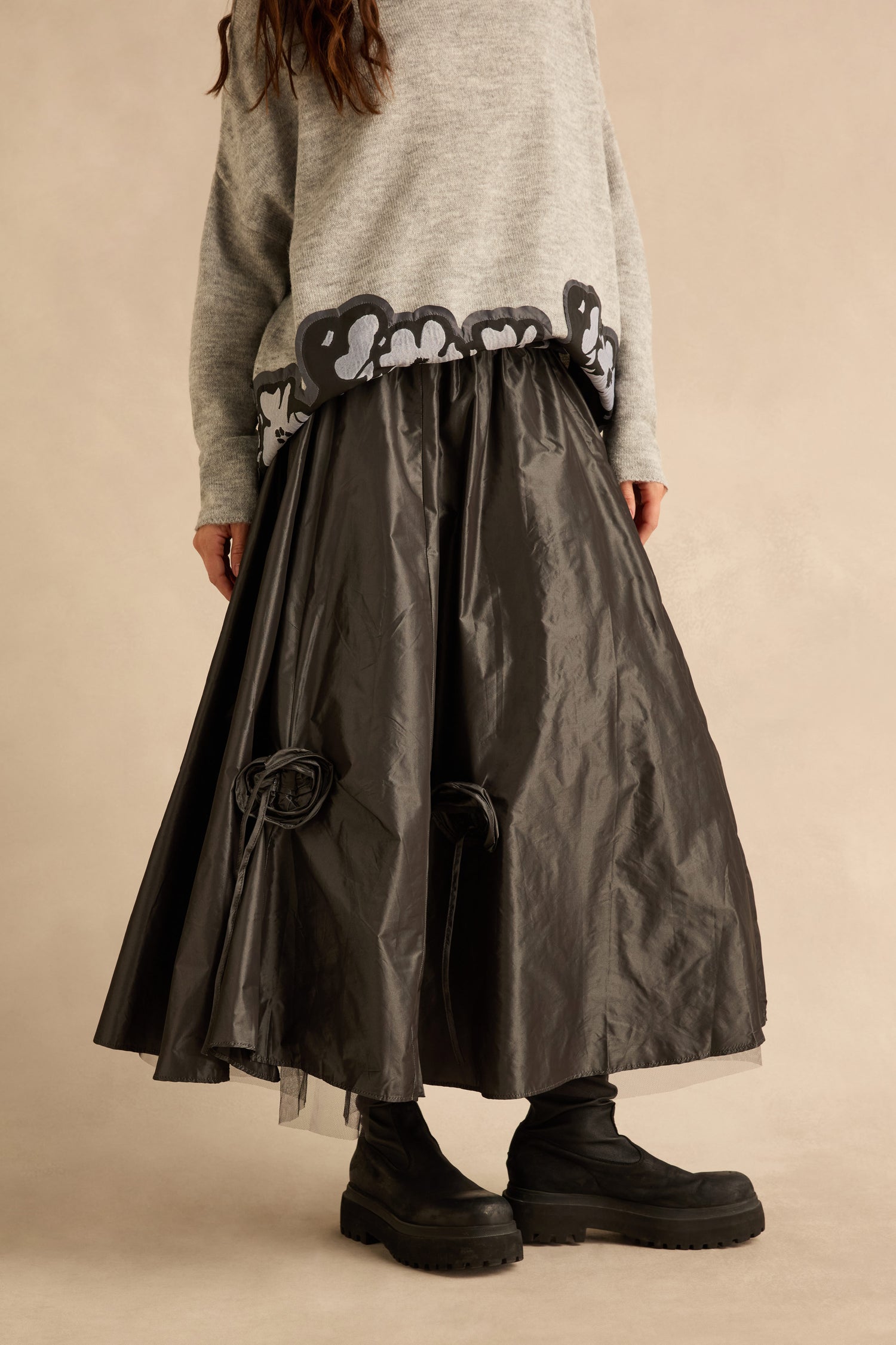 A person wearing a grey sweater, black platform boots, and a Flower Detail Taffeta Skirt, which is a voluminous, calf-length black style with fabric flower accents.