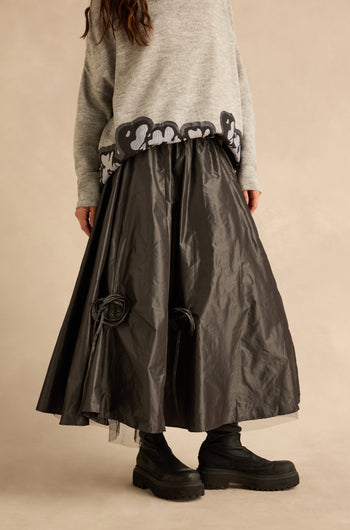 A person wearing a grey sweater, black platform boots, and a Flower Detail Taffeta Skirt, which is a voluminous, calf-length black style with fabric flower accents.