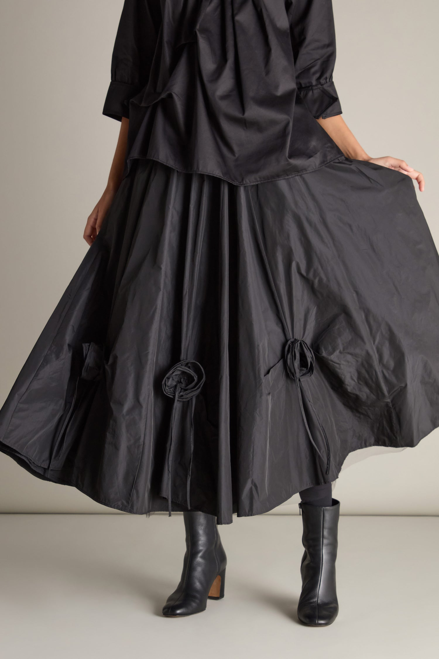 A person wearing the Taffeta Flower Detail Skirt, a long black piece with unique taffeta flower accents, paired with black boots and a loose black top, holds its voluminous silhouette out to the side.