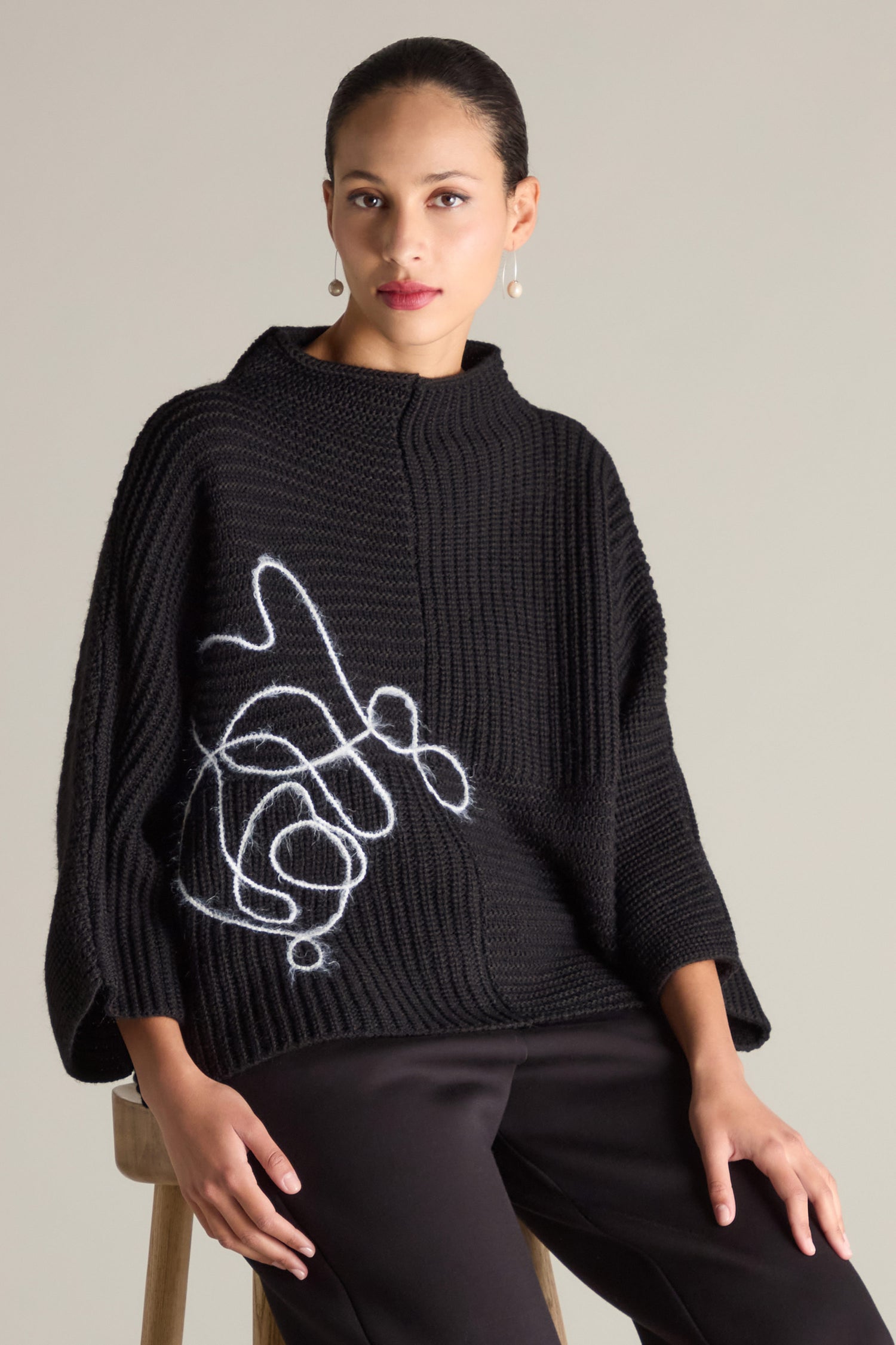 A person wearing the Topstitch Detail Rib Knit in black, featuring a white abstract line design and subtle topstitch details, sits on a wooden stool against a plain background.