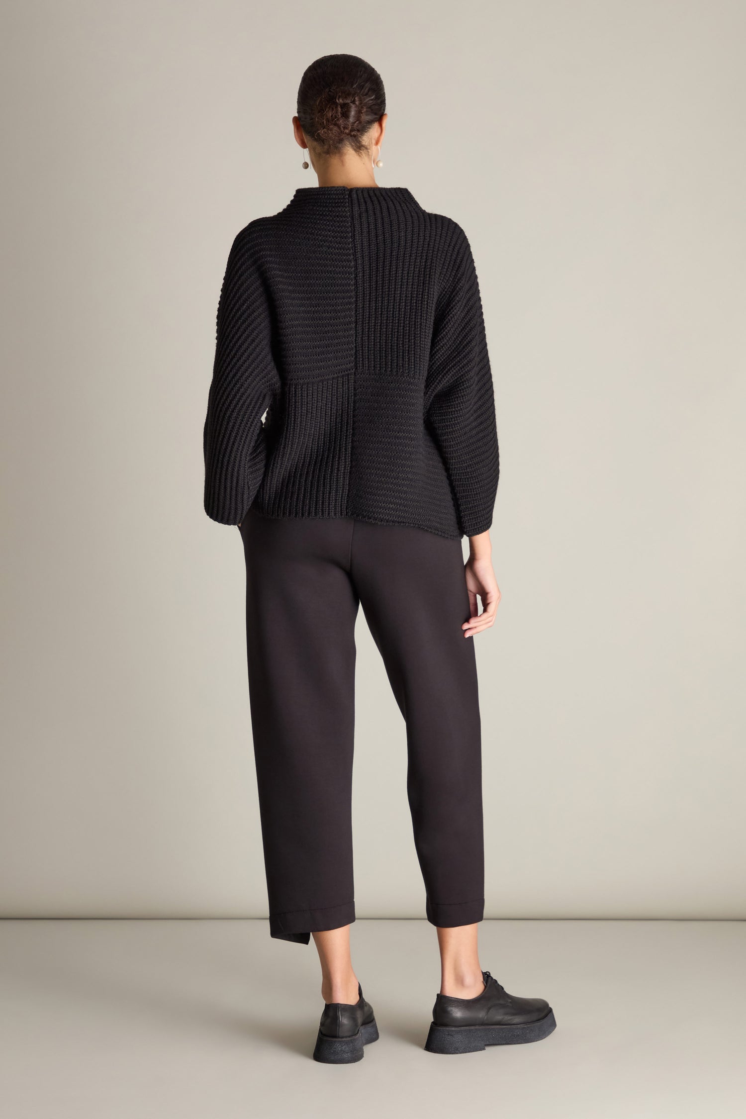 A person dressed in the Topstitch Detail Rib Knit sweater, black pants, and black shoes stands facing away from the camera against a plain background.