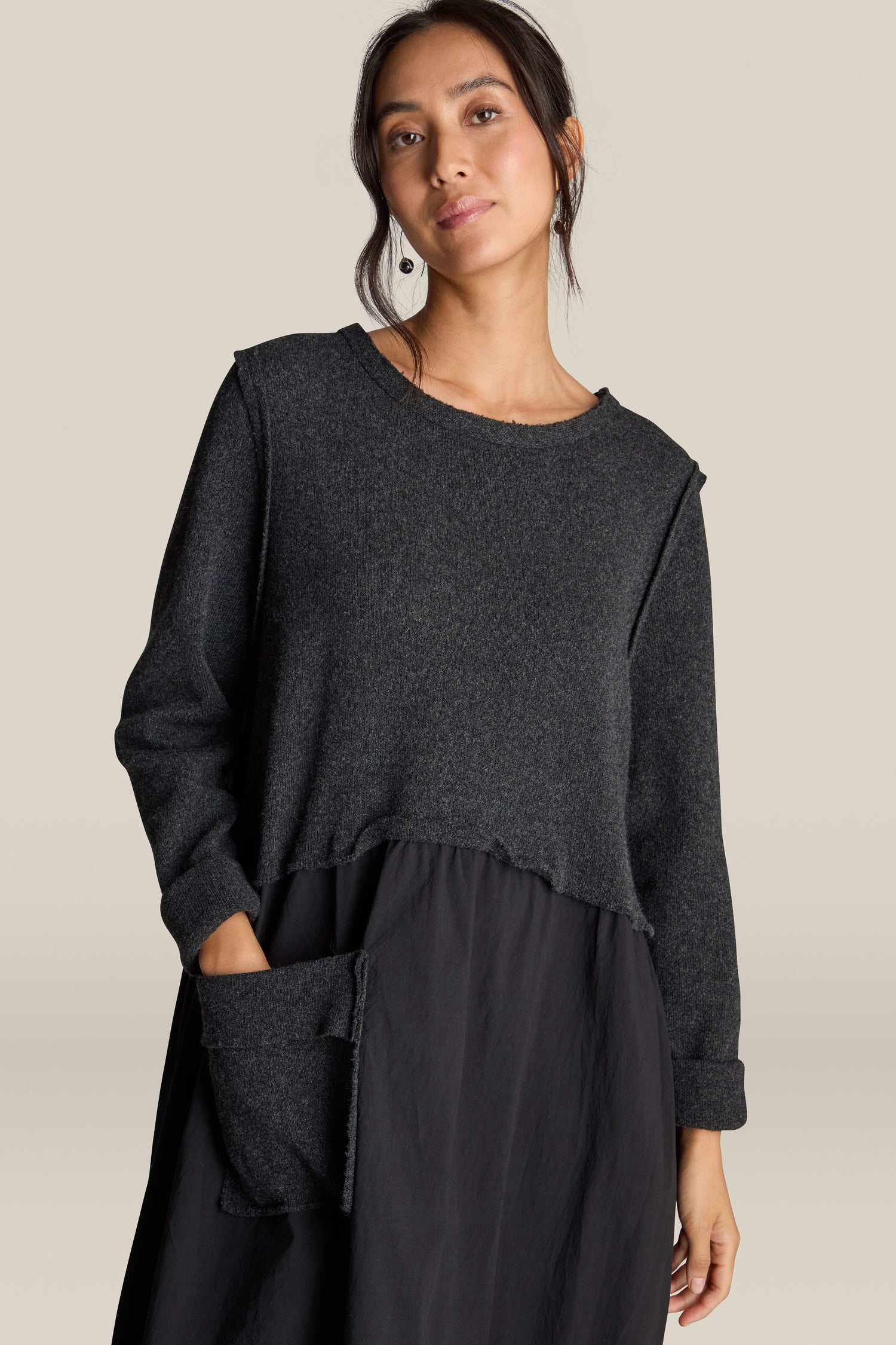 A woman wearing the Patched Easy Dress, a long-sleeved dark gray top featuring patchwork detailing and a front pocket, stands against a plain beige background.