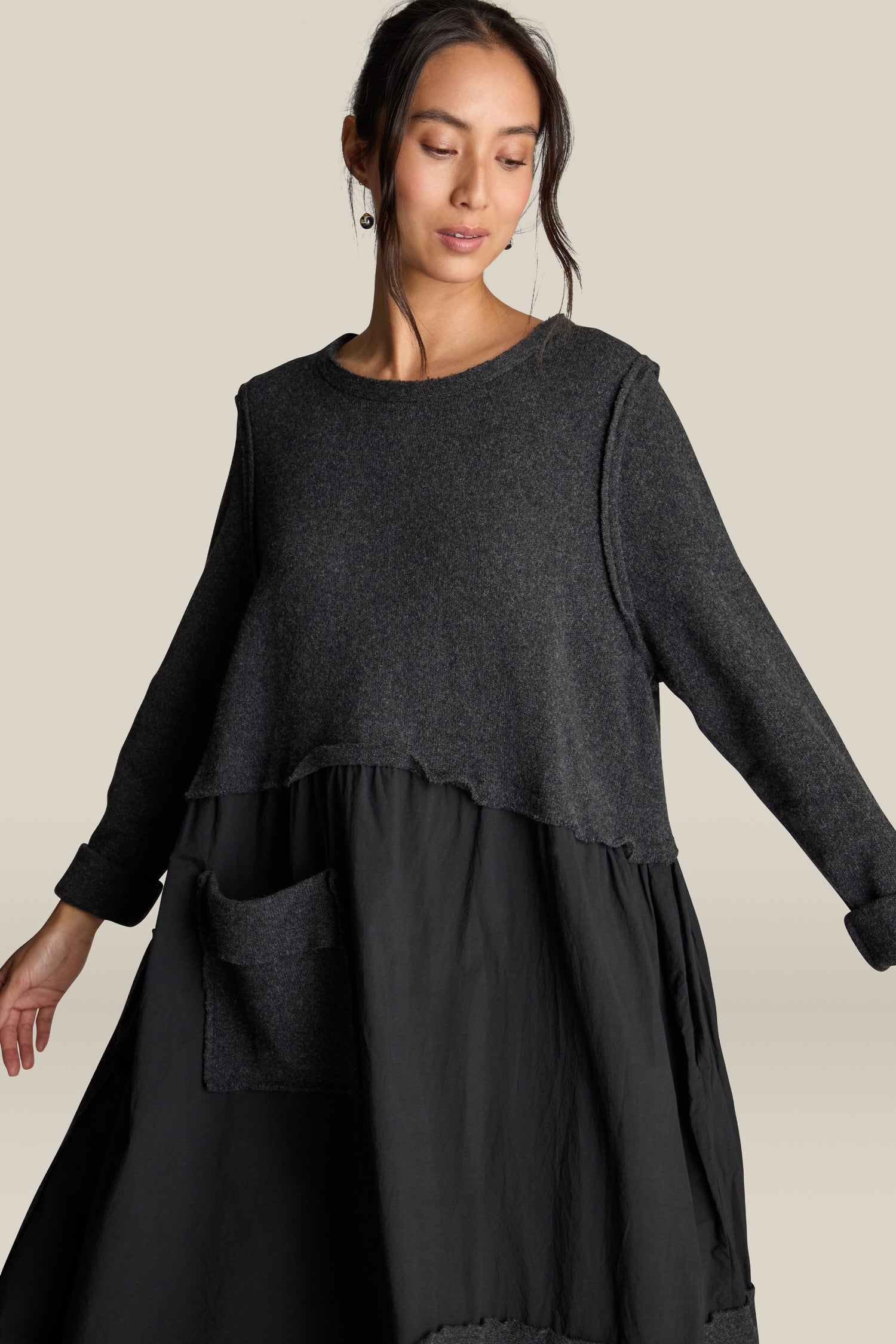 A person wearing the Patched Easy Dress in dark gray, featuring an A-line silhouette with a large front pocket and patchwork detailing, poses against a light background.