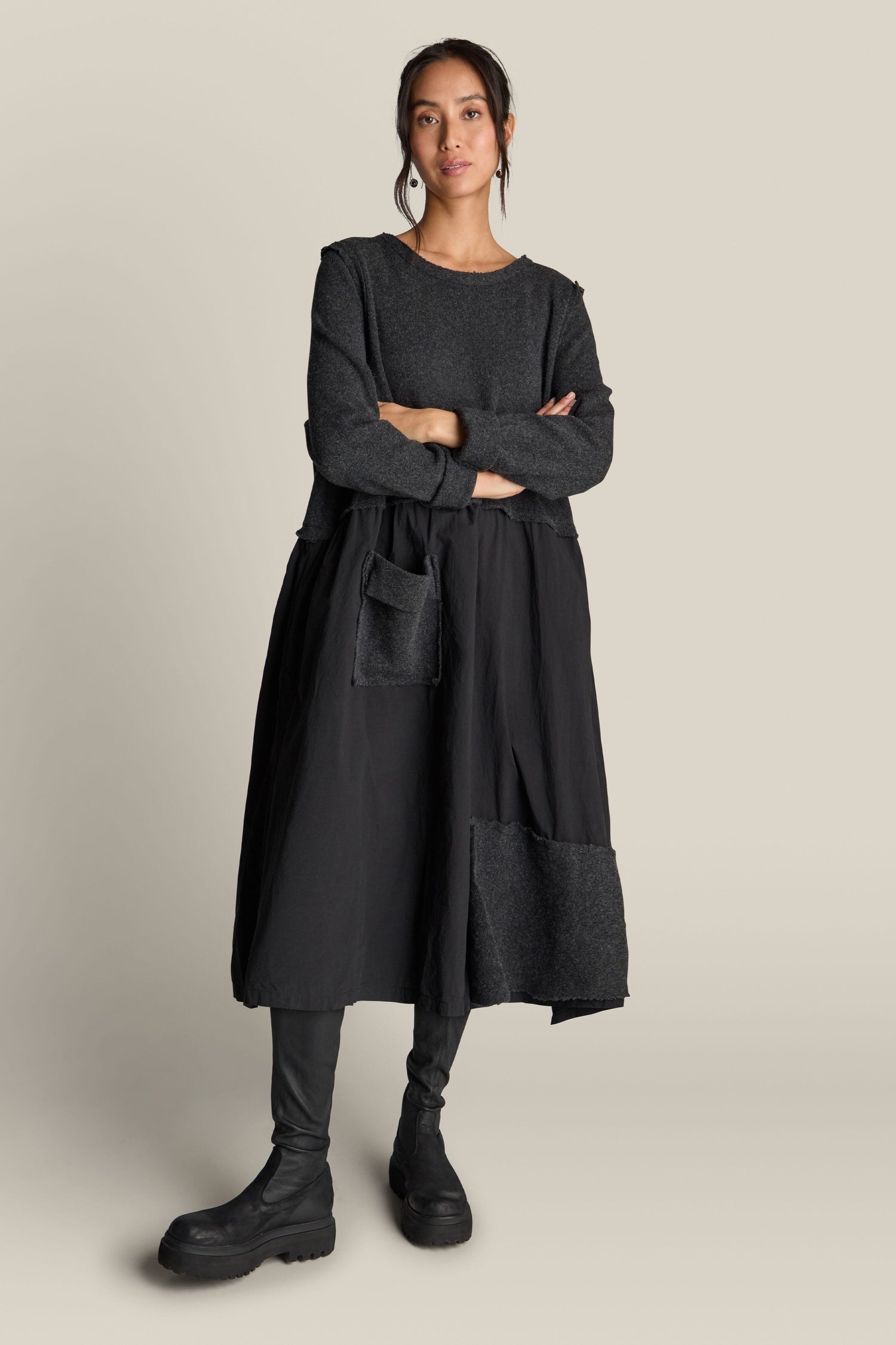 A person stands with arms crossed, wearing a long-sleeved dark sweater, leggings, and bulky black shoes. The outfit is complete with the Patched Easy Dress, characterized by its black design and distinctive patchwork detailing, against a plain background.