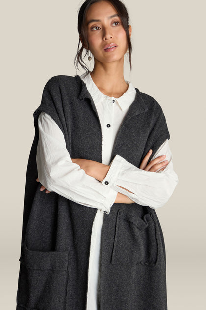 A woman with dark hair is wearing a white shirt and the Long Gilet, a sleeveless long-line dark gray vest with front pockets, crafted from luxurious virgin wool. She stands with her arms crossed, looking slightly off-camera.