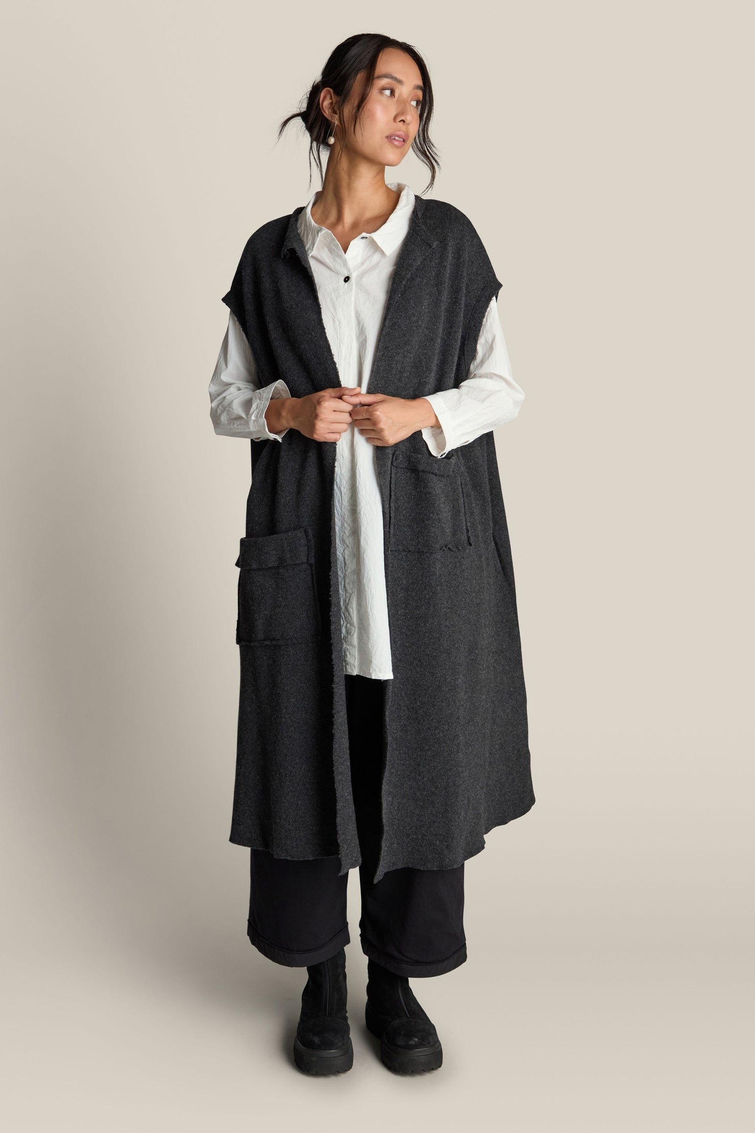 A person stands against a plain background wearing the Long Gilet, a dark gray sleeveless open cardigan made from a luxurious virgin wool blend, over a long white shirt and black pants. They are looking slightly to the side with their hands touching the cardigan.