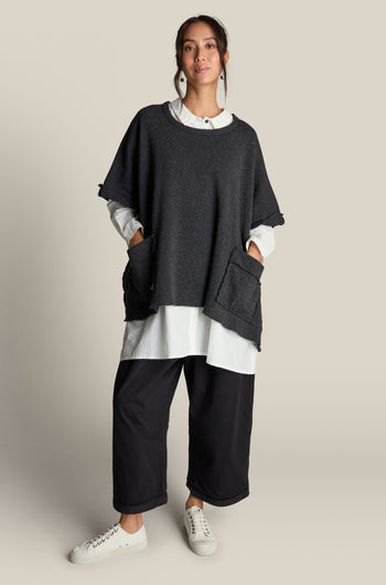 A person stands wearing the Two Pocket Tabard by Barbara Lang, made from a loose dark gray virgin wool blend, layered over a white long-sleeve shirt. They complete their outfit with baggy black pants and white sneakers, accessorized with large earrings while posing against a plain background.