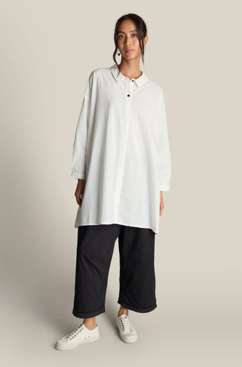 Person wearing a long white button-up shirt, Jersey Turn Up Trousers in black with a wide-leg design, and white sneakers, standing against a plain background.
