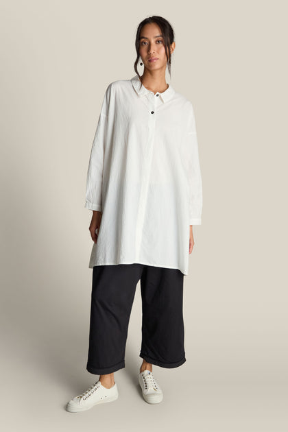 Person wearing a long white button-up shirt, Jersey Turn Up Trousers in black with a wide-leg design, and white sneakers, standing against a plain background.