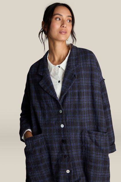 A woman, poised against a plain background, showcases a classic blue and black check pattern coat over a white collared shirt. This Check Jacket, crafted from 100% virgin wool, perfectly merges timeless style with contemporary elegance.