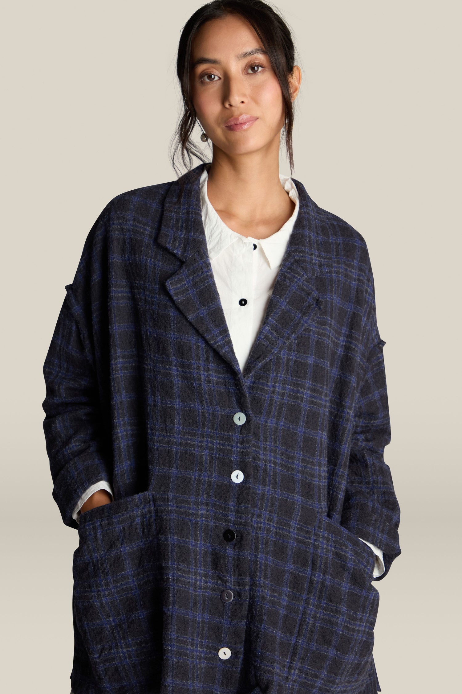 A person wearing the Check Jacket by Barbara Lang, featuring a navy plaid oversized design with white buttons, layered over a white shirt, stands against a plain beige background. Their hands are in the coat pockets.