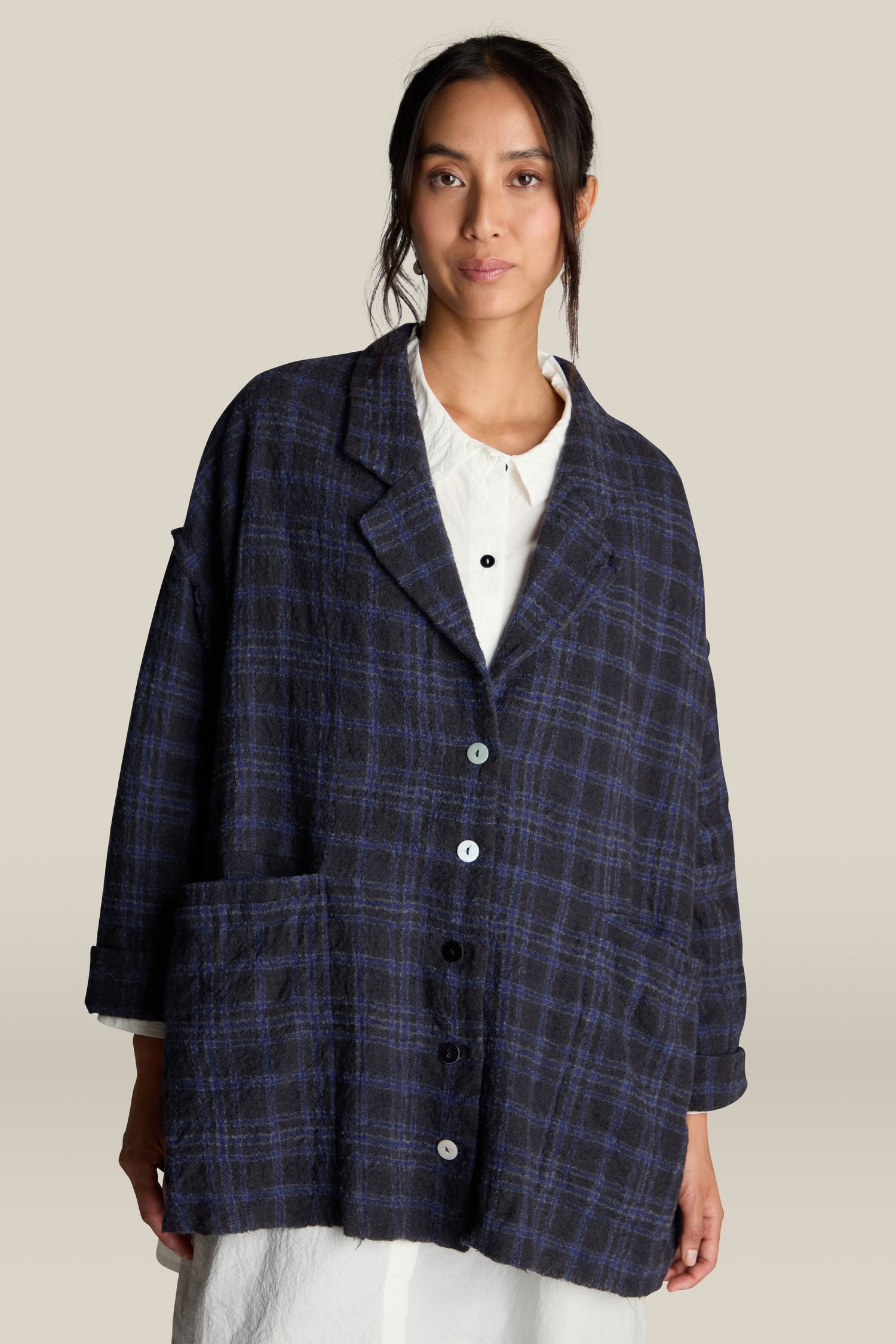 A person is wearing a loose-fitting Check Jacket with large pockets over a white shirt, standing against a plain background. The dark plaid jacket features a classic check pattern and is made from 100% virgin wool.