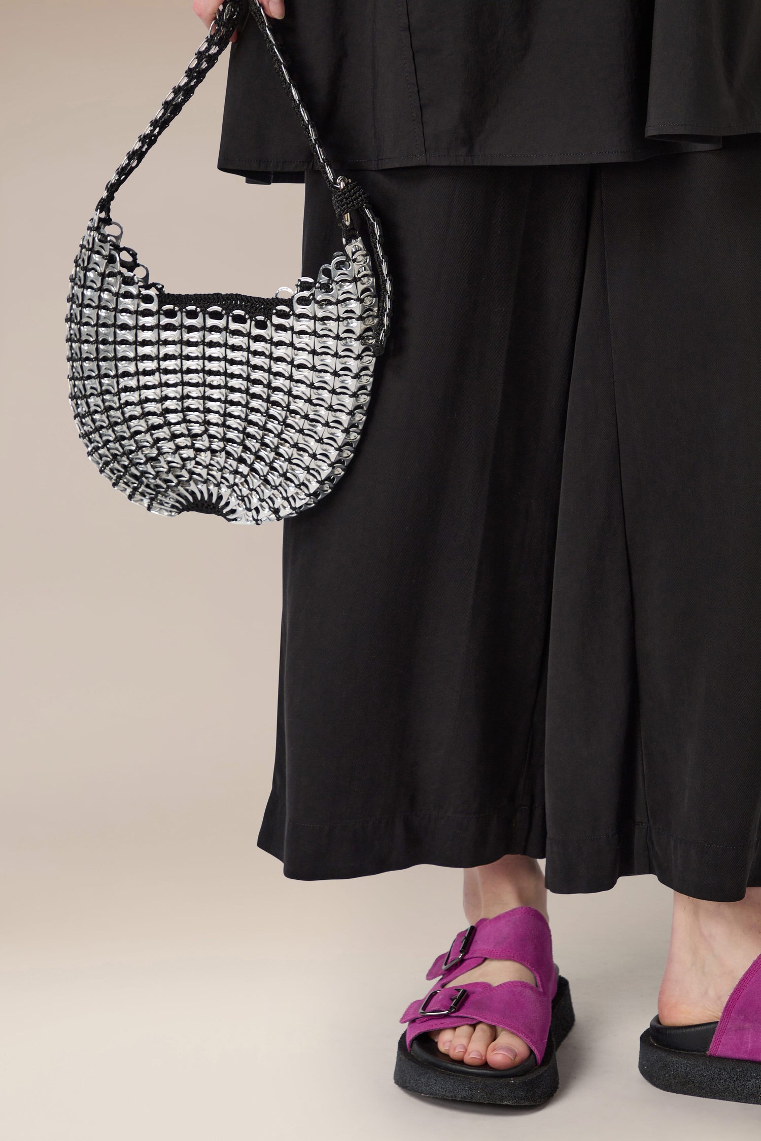 A person wearing black culottes and purple sandals carries a handcrafted in Brazil Bellani Small Bottletop Handbag with its signature chainmail design.